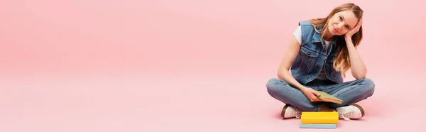 Panoramic shot of young woman in denim clothes sitting on floor near books and propping up head with hand on pink — Stock Photo