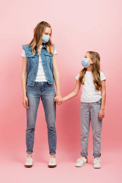 Visão de comprimento total de mãe e filha em jeans e máscaras médicas de mãos dadas e olhando um para o outro em rosa — Fotografia de Stock