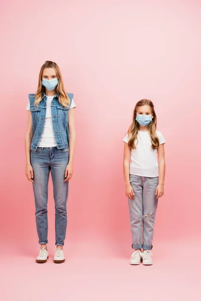 Vista completa de la madre y la hija en máscaras médicas, con ropa de mezclilla, mirando a la cámara en rosa - foto de stock