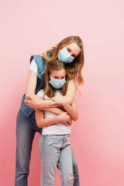 Молодая женщина и ребенок в медицинских масках обнимаются и смотрят на камеру, изолированную на розовый — стоковое фото