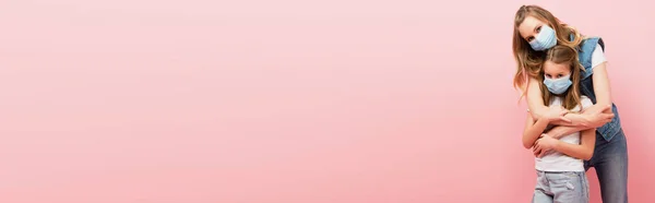 Horizontales Konzept der Frau in Jeanskleidung, die Tochter umarmt, während sie medizinische Masken zusammen isoliert auf rosa trägt — Stockfoto