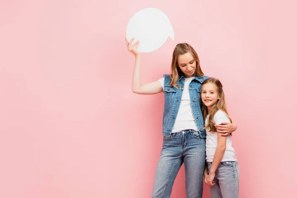 Mujer joven en ropa de mezclilla sosteniendo la burbuja de pensamiento mientras abraza hija aislada en rosa - foto de stock