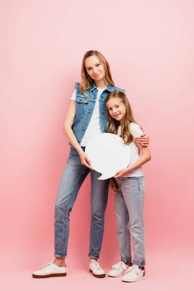 Full length view of young woman in denim vest and jeans embracing daughter while holding thought bubble on pink — Stock Photo