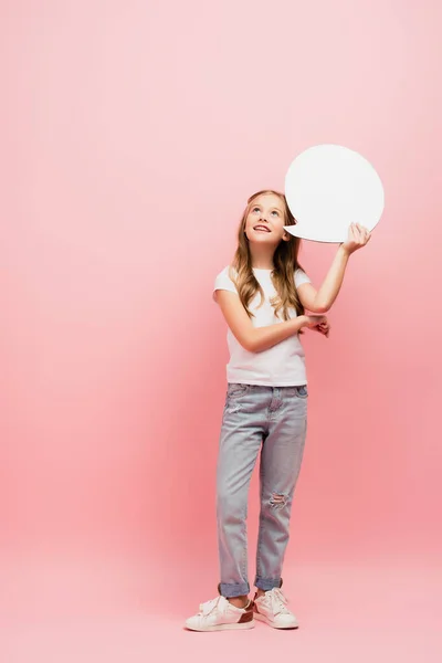 Vista completa de niño en camiseta blanca y pantalones vaqueros azules mirando hacia arriba mientras sostiene la burbuja de pensamiento en rosa - foto de stock