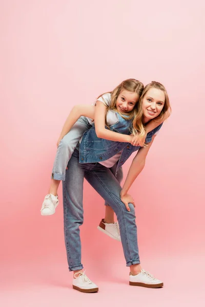 Visão comprimento total da jovem mulher em roupas de ganga piggybacking miúdo em rosa — Fotografia de Stock