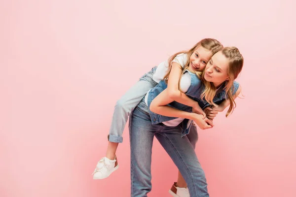 Young woman in denim clothes piggybacking child isolated on pink — Stock Photo