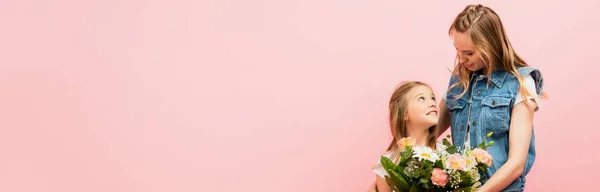 Horizontal concept of young woman presenting bouquet of flowers to daughter while looking at each other isolated on pink — Stock Photo