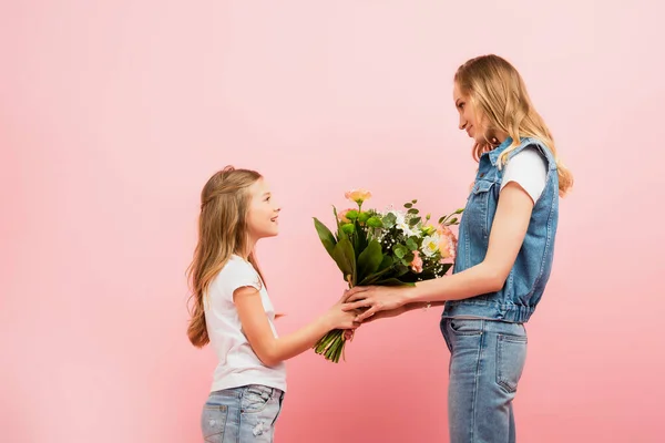 Vista lateral de la niña que presenta ramo de flores a la madre con ropa de mezclilla aislada en rosa - foto de stock