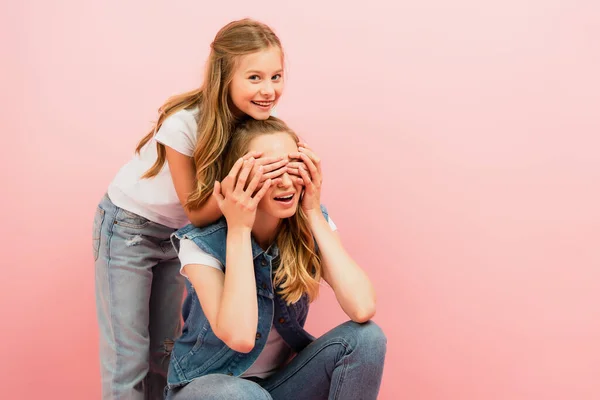Menina olhando para a câmera enquanto cobre mães olhos com as mãos isoladas em rosa — Fotografia de Stock