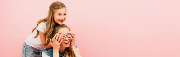 Immagine orizzontale del bambino che copre gli occhi delle madri con le mani e guarda la fotocamera isolata sul rosa — Foto stock
