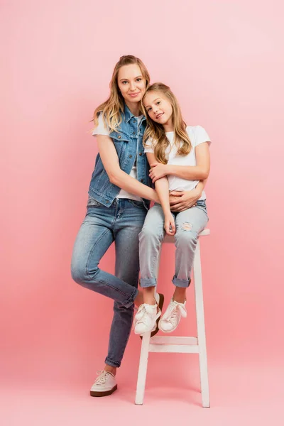 Mãe em roupas jeans abraçando criança sentada em fezes altas e olhando para a câmera em rosa — Fotografia de Stock