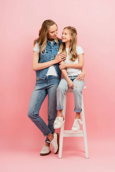 Молодая женщина в джинсовой одежде обнимает дочь сидя на высоком стуле на розовом — стоковое фото