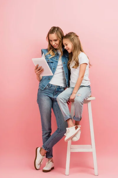 Девушка в джинсах и белой футболке сидит на высоком стуле рядом с матерью, используя цифровой планшет на розовый — стоковое фото