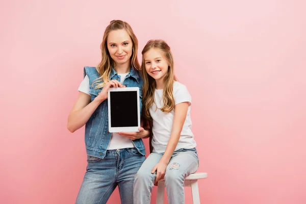 Giovane donna in gilet di denim e jeans con tablet digitale con schermo bianco vicino al bambino seduta su sgabello isolato su rosa — Foto stock