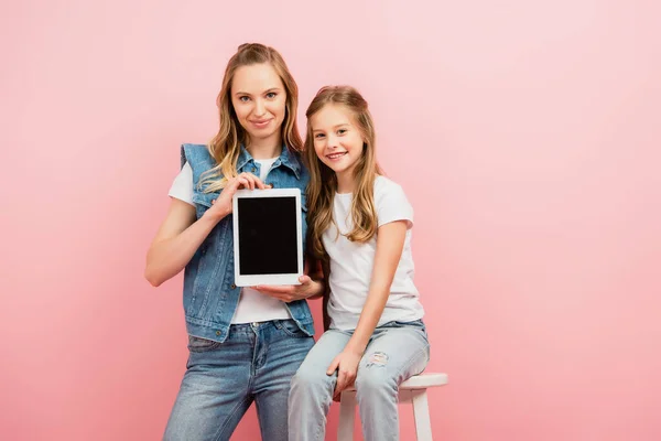 Mujer joven en chaleco vaquero y jeans sosteniendo tableta digital con pantalla en blanco cerca de niño sentado en el taburete aislado en rosa — Stock Photo