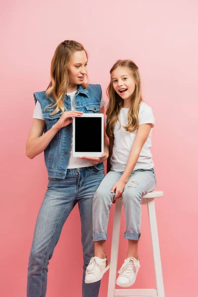 Junge Frau in Jeanskleidung hält digitales Tablet mit leerem Bildschirm in der Nähe aufgeregter Tochter, die auf hohem Schemel sitzt, isoliert auf rosa — Stockfoto