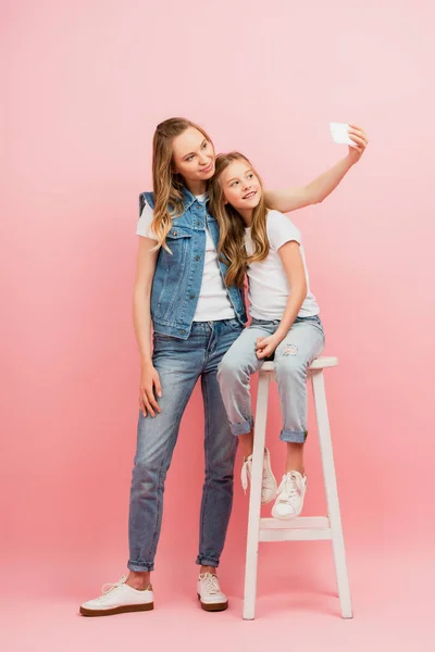 Vue pleine longueur de la jeune femme en vêtements en denim prenant selfie sur smartphone avec sa fille assise sur un tabouret haut sur rose — Photo de stock