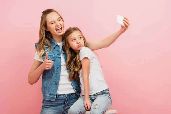 Mujer emocionada en ropa de mezclilla tomando selfie en el teléfono inteligente con la hija haciendo pucheros labios y mostrando el pulgar hacia arriba aislado en rosa - foto de stock