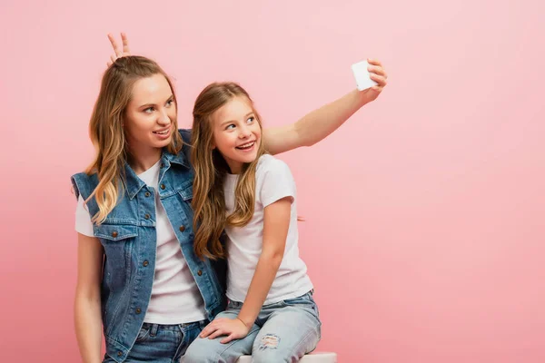 Mädchen zeigt Hasenohren Geste in der Nähe des Kopfes der Mutter, während sie Selfie auf Smartphone auf rosa macht — Stockfoto