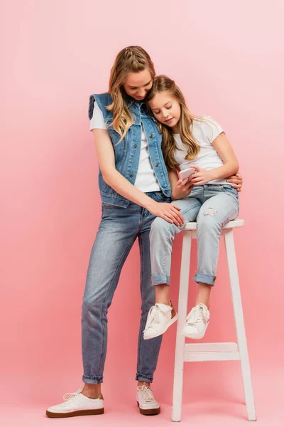 Vue pleine longueur de la femme en jeans et gilet en denim debout près de l'enfant assis sur un tabouret haut et en utilisant un smartphone sur bleu — Photo de stock