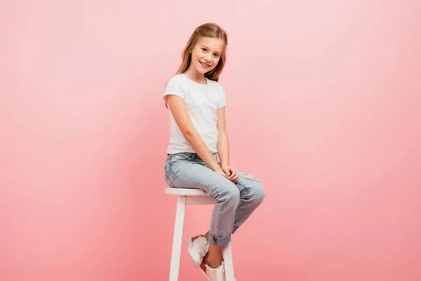 Kind in weißem T-Shirt und Jeans blickt in die Kamera, während es isoliert auf rosa Hochstuhl sitzt — Stockfoto