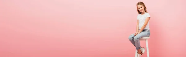 Cabeçalho do site da menina em t-shirt branca e jeans azul sentado em fezes altas e olhando para a câmera isolada em rosa — Fotografia de Stock