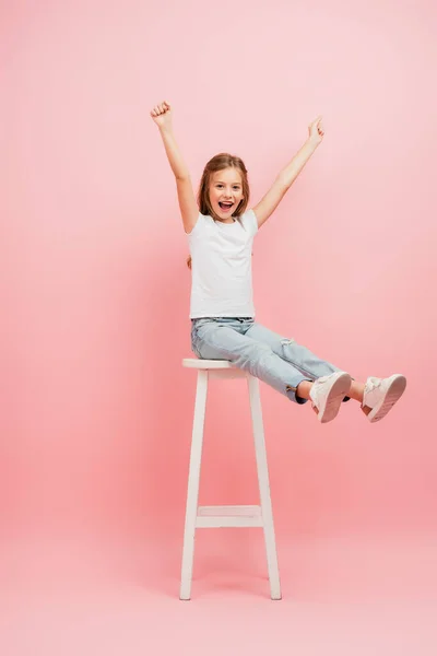 Child in white t-shirt and blue jeans showing winner gesture while sitting on high stool with outstretched legs on pink — Stock Photo