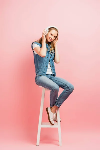 Full length view of woman touching wireless headphones while sitting on high chair and looking at camera on pink — Stock Photo