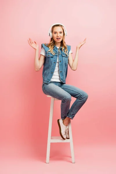Mujer confundida en auriculares inalámbricos que muestran el gesto de encogerse de hombros mientras está sentada en el taburete alto en rosa - foto de stock