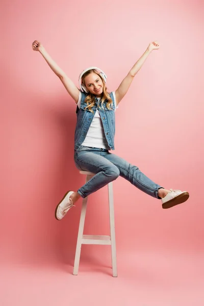 Excited woman in wireless headphones showing winner gesture while sitting on high stool on pink — Stock Photo