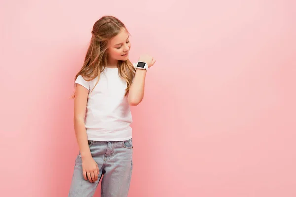 Kid in white t-shirt and jeans looking at smartwatch isolated on pink — Stock Photo