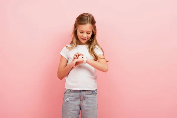 Niño en camiseta blanca y jeans tocando smartwatch aislado en rosa - foto de stock