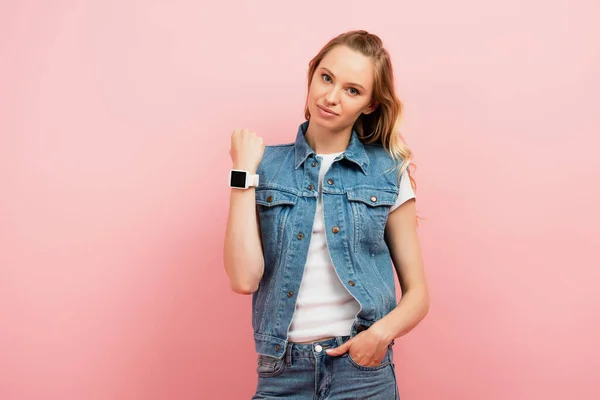 Young woman in denim clothes showing smartwatch on wrist while standing with hand in pocket isolated on pink — Stock Photo
