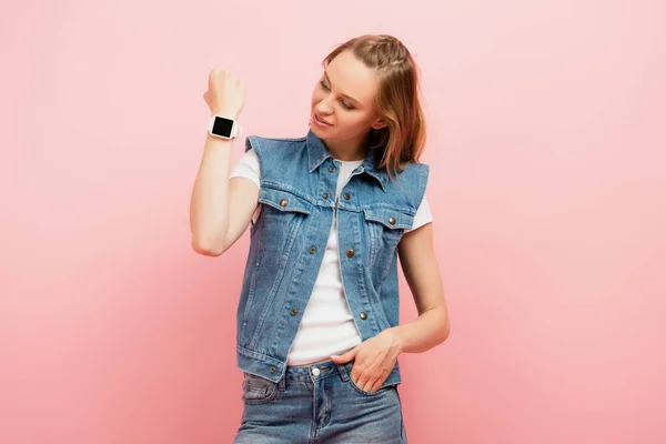 Mujer joven en chaleco vaquero que muestra smartwatch en la muñeca mientras está de pie con la mano en el bolsillo aislado en rosa - foto de stock