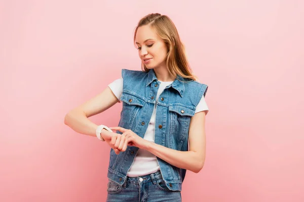 Jovem mulher em colete jeans tocando smartwatch isolado em rosa — Fotografia de Stock