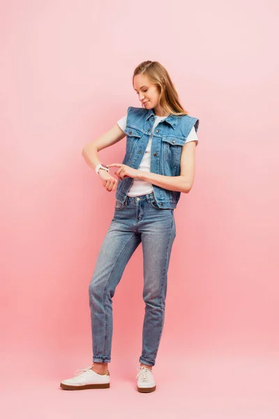 Повний вигляд молодої жінки в джинсовому одязі, що торкається розумного годинника на рожевому — стокове фото
