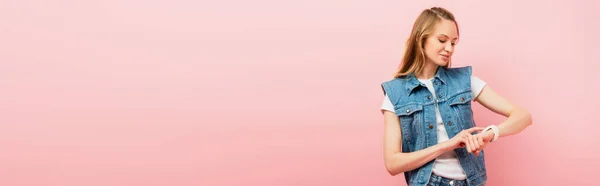 Панорамный снимок молодой женщины в джинсовой одежде, касающейся умных часов, изолированных на розовом — стоковое фото