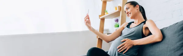 Панорамний знімок вагітної жінки в спортивному одязі, що приймає селфі зі смартфоном на дивані — стокове фото