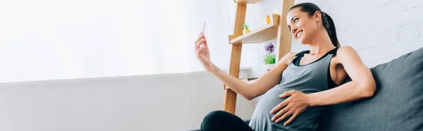 Prise de vue panoramique de la femme enceinte en tenue de sport prenant selfie avec smartphone sur canapé — Photo de stock