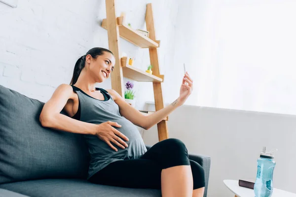 Sportiva incinta che prende selfie con smartphone vicino alla bottiglia sportiva — Foto stock