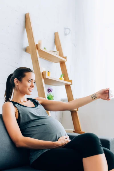 Enfoque selectivo de la joven embarazada en ropa deportiva tomando selfie con teléfono inteligente en el sofá - foto de stock