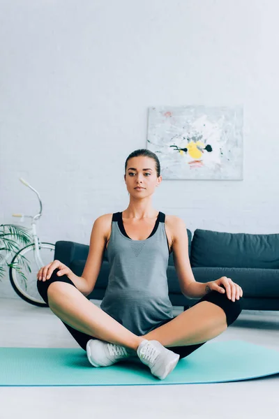 Deportista embarazada sentada con piernas cruzadas en postura de yoga en colchoneta de fitness en casa - foto de stock
