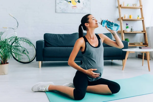 Donna incinta che beve acqua durante l'allenamento sul tappeto fitness a casa — Foto stock
