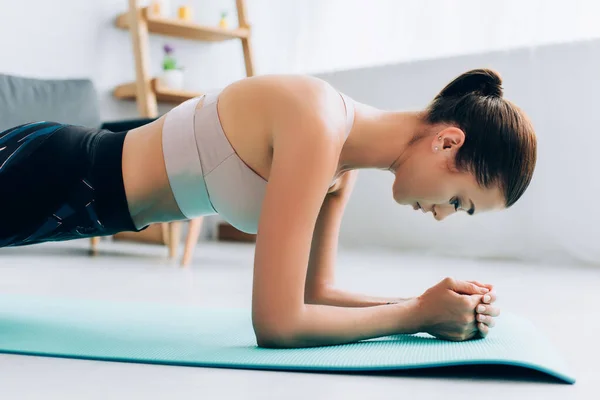 Seitenansicht einer brünetten Frau, die in Planke steht, während sie zu Hause trainiert — Stockfoto
