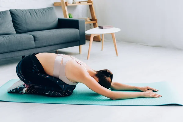 Seitenansicht der brünetten Sportlerin, die sich dehnt, während sie im Wohnzimmer auf einer Fitnessmatte sitzt — Stockfoto