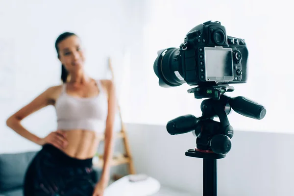 Selective focus of digital camera on tripod beside fit sportswoman in living room — Stock Photo