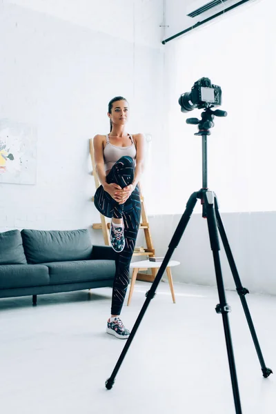 Selective focus of brunette sportswoman warming up before training near digital camera at home — Stock Photo