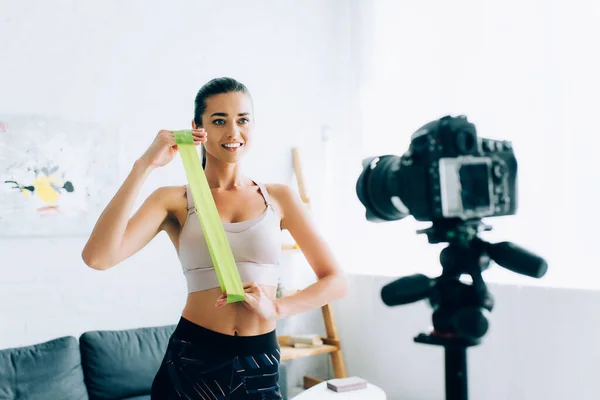 Selective focus of sportswoman holding resistance band near digital camera on tripod at home — Stock Photo