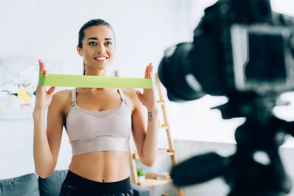 Selective focus of fit sportswoman holding resistance band near digital camera in living room — Stock Photo