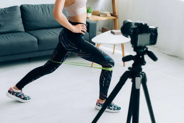 Cropped view of sportswoman using resistance band while doing lunges near digital camera at home — Stock Photo